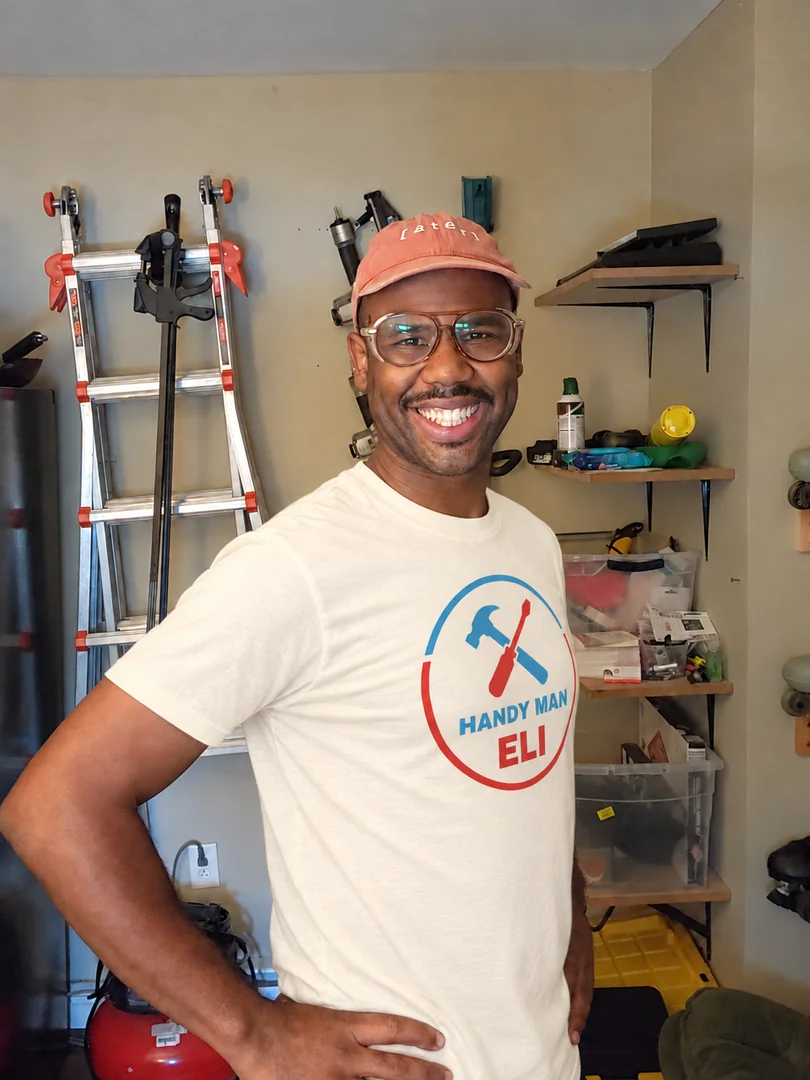 Eli smiling in his workshop, wearing a Handy Man Eli shirt